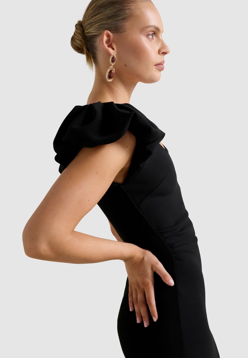 Profiel van een vrouw met haar in een knot, gekleed in een zwarte jurk met grote gerimpelde schouders en goudbruin hang oorbellen, hand op haar onderrug.