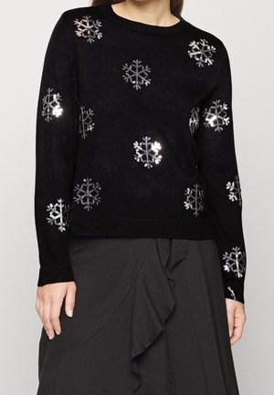 Pull noir à manches longues avec des flocons de neige en sequins argentés, porté avec une jupe noire à volants.