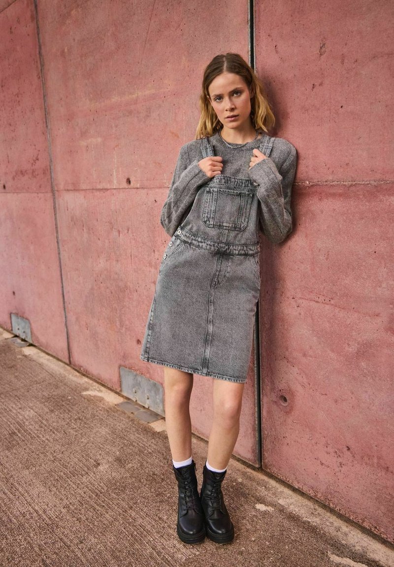 Grå denim overallklänning med en framficka, tillsammans med en grå sweater och svarta snörboots. Outfiten har en ren och enkel design.