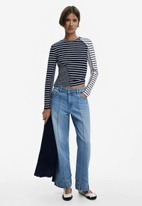 Langärmeliges gestreiftes Oberteil in Marineblau und Weiß, kombiniert mit weit geschnittenen hellblauen Jeans mit markanten Nähten und ausgestellten Säumen. Schwarz-weiße Schuhe.