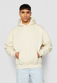 Beige hettegenser laget av mykt stoff, med kengurulomme, ribbede mansjetter og en subtil logo på brystet. Brukt med lyseblå jeans.