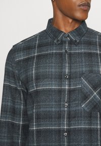 Homme portant une chemise à carreaux gris foncé et noir avec des boutons, une poche avant et un col boutonné, recadré sous le nez.