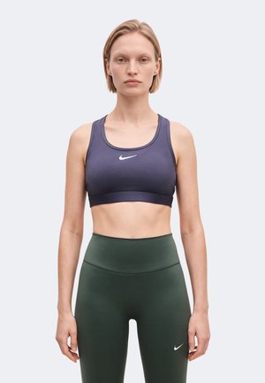 Une femme porte un soutien-gorge de sport Nike violet et un legging vert foncé taille haute sur un fond clair et uni.