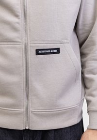 Haut à capuche zippé gris clair avec une texture douce, arborant un patch logo noir "REDEFINED REBEL" sur la poitrine et des poches latérales.