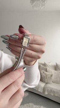 Mains aux ongles argentés métalliques tenant une petite montre argentée avec un bracelet orné de strass, sur un fond doux de salon.