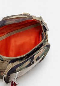 Borsa con motivo mimetico, dotata di parte superiore con zip, rivestimento interno arancione e una piccola tasca esterna. Presenta spallacci regolabili e accessori in argento.