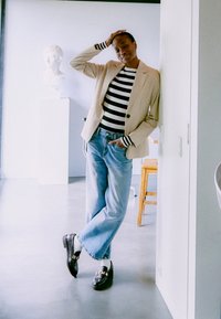 Bežinis švarkas virš juodos ir baltos smulkaus dryžių marškinėlių, šviesiai mėlynos pločio kojinės džinsai, juodi batai su auksinėmis detalėmis ir baltos kojinės. Minimalistinė fono scena.