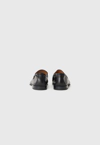 Une paire de mocassins en cuir noir montrée de l'arrière sur un fond blanc uni, avec de petits talons empilés et des bords arrondis.