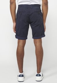 Man in marineblauwe opgerolde shorts, witte sneakers en een wit shirt, van achteren afgebeeld tegen een eenvoudige lichte achtergrond.