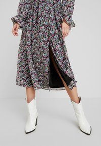 Femme portant une robe midi fleurie avec une fente latérale et des manches longues, associée à des bottines blanches à bout pointu et talons carrés.
