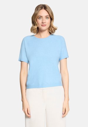 Kobieta o blond włosach do ramion, ubrana w jasnoniebieski sweter z krótkim rękawem i białe spodnie, stojąca na jednolitym tle.