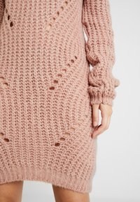 Robe tricotée rose claire avec un tissage lâche et texturé, présentant des détails ajourés et des manches longues, se terminant au-dessus du genou.