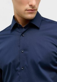Man in een donkerblauw overhemdd met puntsluiting, weergegeven van kin tot borst tegen een effen achtergrond.