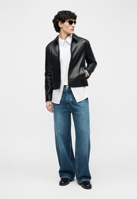 Άνδρας που φορά μαύρο δερμάτινο μπουφάν, λευκό πουκάμισο με κουμπιά, φαρδιά μπλε τζιν, μαύρα παπούτσια και γυαλιά ηλίου, στέκεται με το ένα χέρι στην τσέπη.