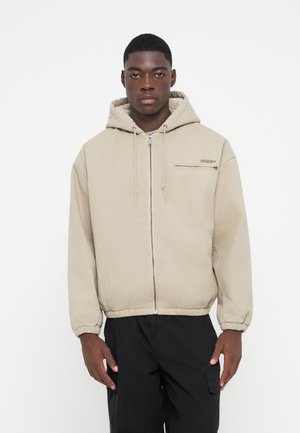 Beige hooded jacket with a zip front, elastic cuffs, and a small chest pocket. Textured fabric with a soft inner lining. Paired with black pants.