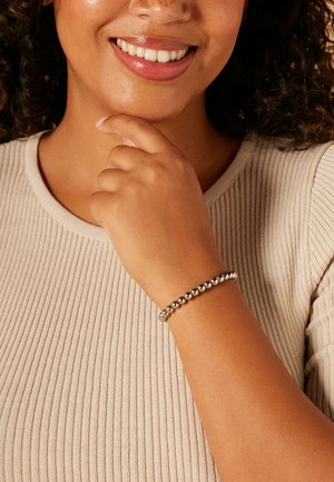 Silbernes Perlenarmband mit glatten, runden Metallperlen auf einem schlanken elastischen Band, getragen an einem Handgelenk vor einem strukturierten beigen Oberteil.