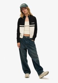 Veste aviateur à fermeture éclair noire et crème avec deux bandes, associée à un haut court blanc et un jean ample foncé. Casquette verte avec le logo "SUPERDRY".