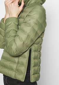 Olivově zelená puffer bunda s horizontálním prošíváním, se zipem na boku a vysokým límcem. Vyrobena z lehkého, lesklého materiálu.