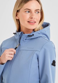 Light blue softshell jacket with a zip closure, adjustable hood, and black hardware. Features a logo patch on the left sleeve.