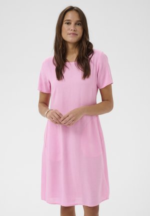 Femme aux longs cheveux bruns portant une robe rose à manches courtes, longueur genou, avec un col rond et des poches latérales, sur fond blanc uni.