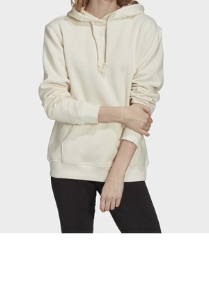 Personne portant un sweat à capuche couleur crème avec poche avant et pantalon noir, debout avec un bras croisé et l'autre pendant.