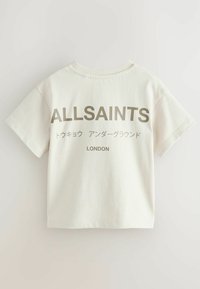 Krémové tričko s krátkým rukávem a kulatým výstřihem, se silným nápisem "ALLSAINTS" a dalším stylizovaným textem v japonštině, umístěným uprostřed na zadní straně.