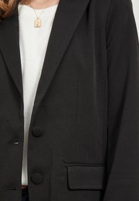 Blazer noir à texture lisse, fermeture à un bouton et revers crantés. Comprend une poche latérale et une coupe cintrée.