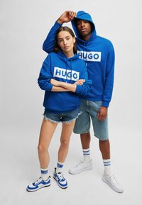 Blue hoodies with white "HUGO" logo; model on left in shorts; both wearing blue and white sneakers and socks. Minimalist design.