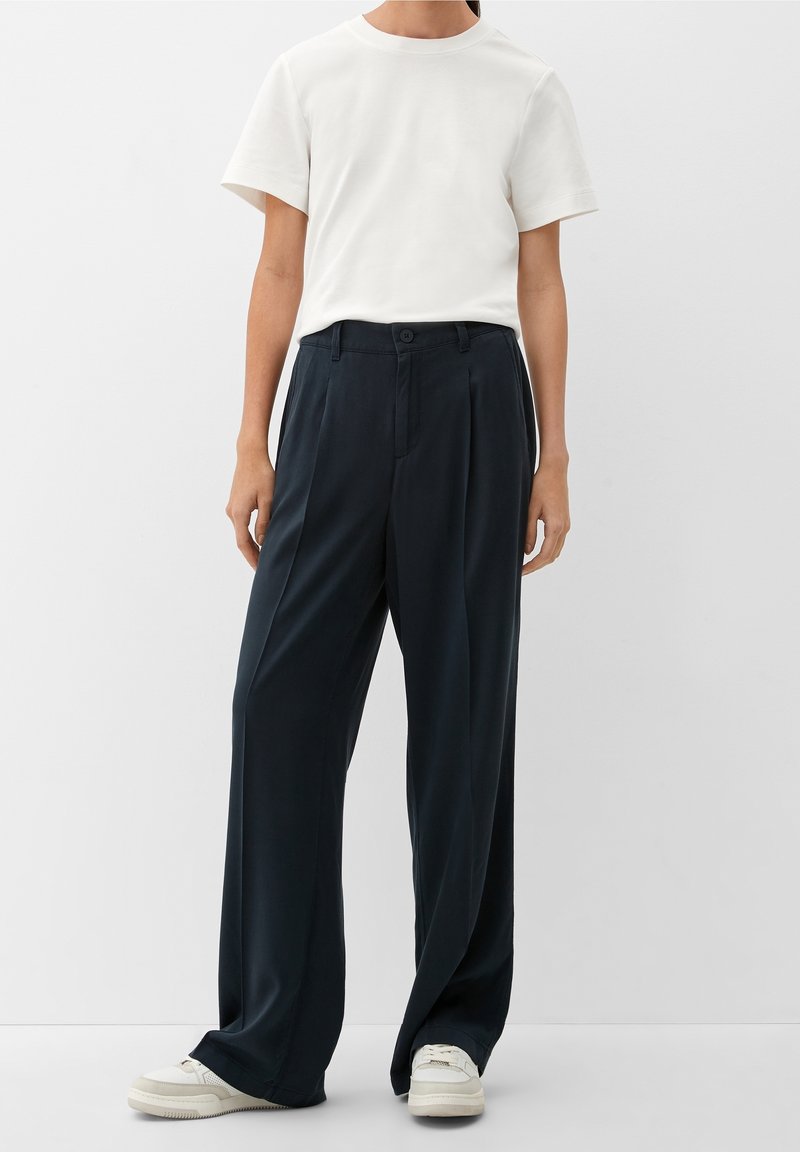 Personne portant un t-shirt blanc uni à manches courtes, un pantalon noir ample taille haute et des baskets blanches, debout devant un fond uni.