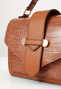 Ruskea krokotiilikaavion nahkalaukku, jossa on muotoiltu rakenne, kultasävyinen kiinnitysklipsi ja litteä olkahihna.