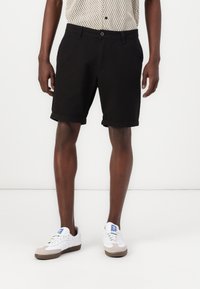 Svarta bomullsshorts med uppvikta fållar, med sidofickor och knäppning, ihop med vita skor och en mönstrad skjorta.