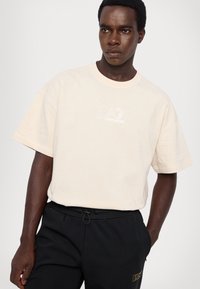 T-shirt beige clair à manches courtes avec un col rond et un logo brodé "EA7 Emporio Armani". Fabriqué en coton doux.