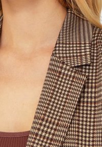 Bruine en crèmekleurige geruite blazer met een reverskraag en gestructureerde schouders; de stof lijkt textuurrijk en van middelzware kwaliteit.