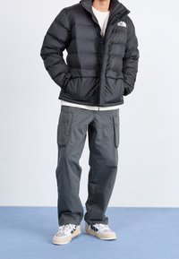 Giacca a vento nera con design trapuntato, zip e tasche laterali, abbinata a pantaloni cargo grigi e sneakers bianche.