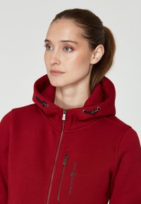 Sudadera roja con cremallera y cuello alto, con cordones y herrajes en negro. Cuenta con un bolsillo en el pecho con cremallera y un logo bordado de "SAIL RACING".