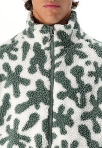 Veste en polaire zippée avec une base blanche et des motifs abstraits verts. Col montant et surface texturée avec des détails de fermeture éclair contrastés.