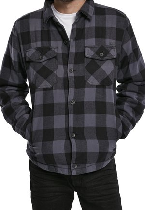 Homme portant une chemise en flanelle à carreaux noirs et gris, boutonnée, avec deux poches poitrine, par-dessus un T-shirt blanc, mains dans les poches.