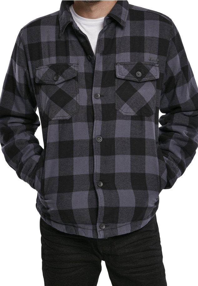 Homme portant une chemise en flanelle à carreaux noirs et gris, boutonnée, avec deux poches poitrine, par-dessus un T-shirt blanc, mains dans les poches.