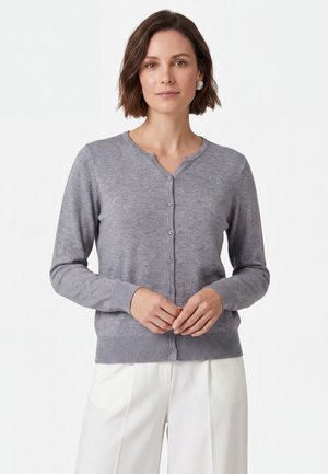 Kvinde med kort brunt hår iført en grå knappet cardigan og hvide bukser, står mod en ensfarvet hvid baggrund.