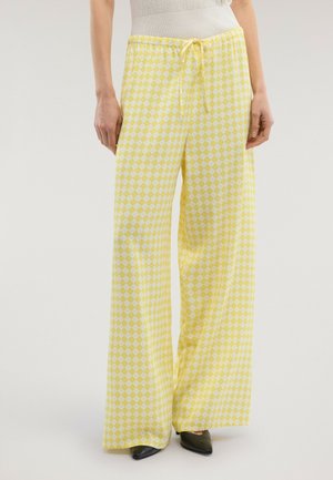 Kvinde iført gule og hvide ternede bukser med høj talje og vide ben, strammet med snøre i taljen, samt sorte spidse sko.