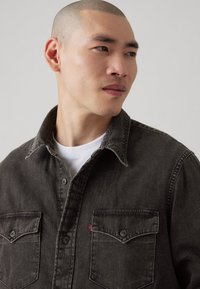 Cămașă din denim gri închis, cu închidere cu nasturi în față, două buzunare pe piept și cusături contrastante. Design cu guler și textură netedă.