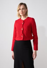 Veste courte texturée rouge avec des manches longues, un col rond, une fermeture à boutons dorés et deux poches avant. Portée sur une jupe noire.
