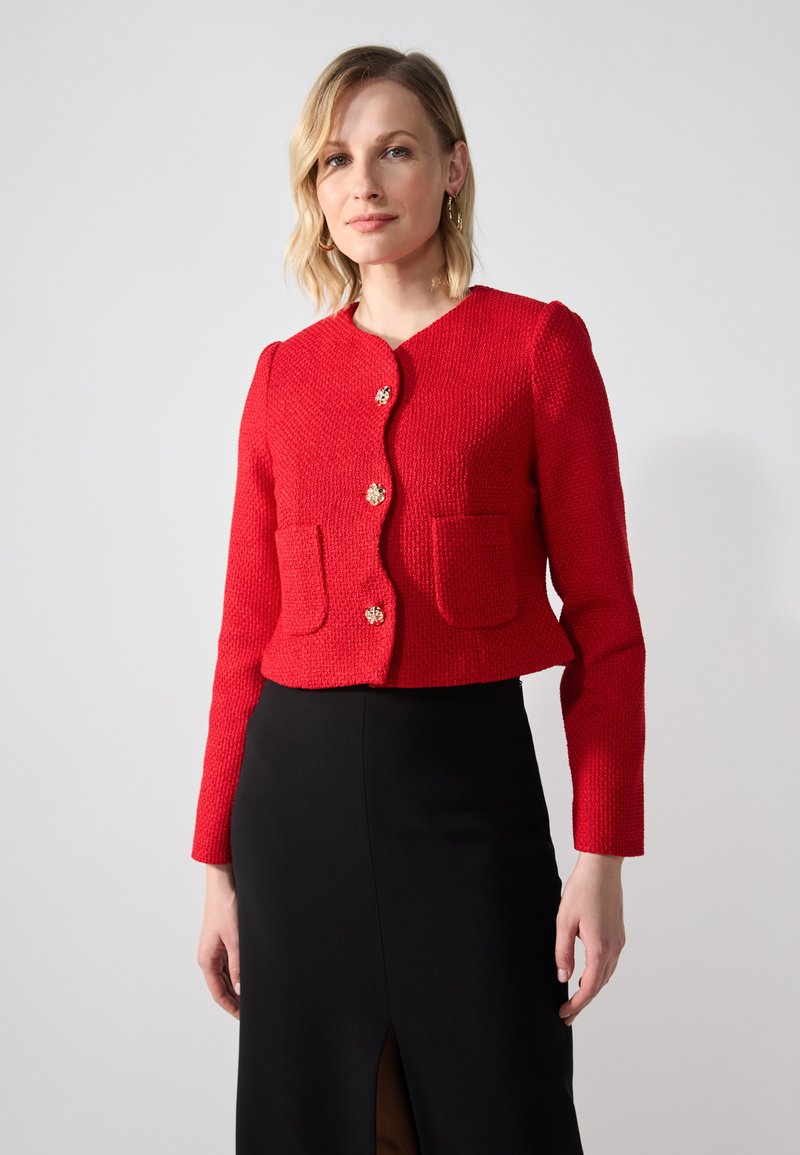 Veste courte texturée rouge avec des manches longues, un col rond, une fermeture à boutons dorés et deux poches avant. Portée sur une jupe noire.