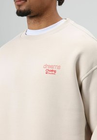 Beige sweater met geribbelde ronde hals, met roze geborduurde tekst "dreams Chasing NOT deadlines." Soepele textuur, losse pasvorm.