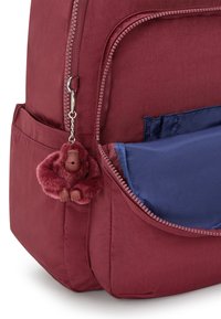 Vinröd ryggsäck med framficka delvis öppen som visar blått foder, och en vinröd plyschgorilla nyckelring hängande från dragkedjan.