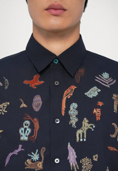 Marineblå skjorte med knapper som har flerfargede broderte motiver av dyr og planter, med en matchende turkis knapp ved kragen.