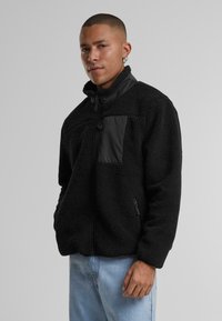 Jachetă din fleece neagră cu fermoar și guler înalt, având un panou negru neted pe piept și două buzunare laterale cu fermoar.