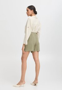 Helle beige Langarmbluse, lässiger Schnitt; olivgrüne Shorts mit elastischem Bund; cremefarbene Mules mit spitzem Zeh und niedrigem Absatz.