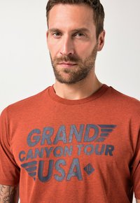 Červené bavlněné tričko s velkým grafickým potiskem "GRAND CANYON TOUR USA" v tmavě modré barvě s křídlo podobnými akcenty.