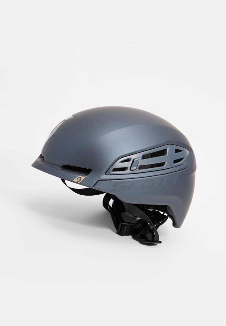 Šedá cyklistická přilba s ventilačními otvory, matným povrchem, nastavitelným popruhem a výrazným logem značky na boku.